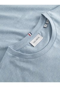 Šviesiai mėlyna T-marškiniai, pagaminti iš minkštos medžiagos, su apvaliu iškirpimu ir logotipo etikete. Apykaklė turi raudoną ir mėlyną akcentą.