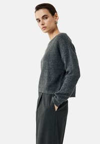 Grauer ribbeliger Strickpullover mit Rundhalsausschnitt und langen Ärmeln, kombiniert mit hochgeschnittenen dunklen Hosen. Glatte Textur, lässige Passform.