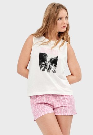 Femme aux longs cheveux blonds portant un débardeur blanc Beatles Abbey Road et un short rose orné du texte ""The Beatles"" en répétition, debout devant un fond uni.