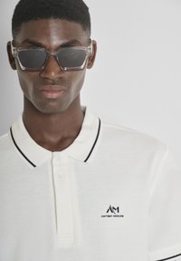 Polo-shirt beige clair avec des finitions noires et un logo sur la poitrine gauche ; associé à des lunettes de soleil transparentes. Tissu lisse et design ajusté.