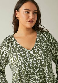 Grünes gemustertes Kleid mit V-Ausschnitt, aus strukturiertem Stoff mit weißen Blumenmotiven. Lange Ärmel mit lockerer Passform.