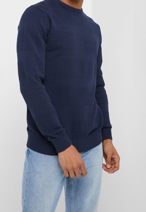 Homme portant un pull bleu marine en maille texturée avec des motifs horizontaux et un jean bleu clair, debout devant un fond clair uni.