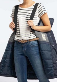 Chaqueta de plumón con exterior azul marino y forro gris, que presenta correas negras. Llevada sobre una camiseta de rayas, con jeans azules y un cinturón marrón.
