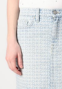 Main gauche reposant à côté d'une jupe en denim bleu avec un motif répété du logo DKNY bleu clair, avec un bouton et une poche visibles.