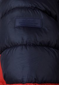Marinblå puffjacka med röd nederdel, med en texturerad yta och en framträdande pr präglad etikett som säger "minoti ETABLERAD ÅR 1997."