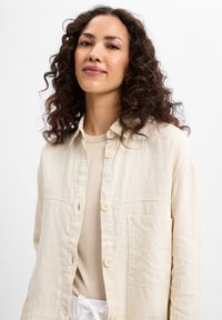 Beige linen shirt with button closure, long sleeves, and a chest pocket, featuring a soft texture and relaxed fit.