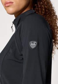 Juodas sportinis švarkas su aukštu apykakle, užtrauktuku ir EA7 logotipu dešiniajame rankovėje. Švelni, struktūruota medžiaga.