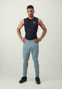 Črna brezrokavnica za šport, svetlo modri usnjeni jogger hlači z oranžnimi stranskimi črtami, stoji na nevtralni podlagi, oblečen v črne športne čevlje.