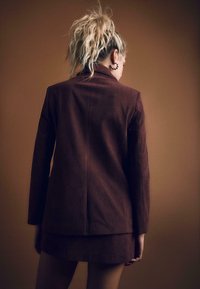 Ruskea corduroy-bleiseri ja minihame-setti, jossa on ribattu pinta ja siistit linjat. Takaviistosta näkyy istuva muotoilu.