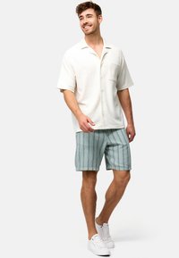 Chemise crème à manches courtes avec boutonnière et poche, associée à un short bleu clair à rayures et des baskets blanches. Tissu simple et texturé.