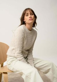 Jasnoszary sweter z splotem warkocza, okrągłym dekoltem i ściągaczem na dole, zestawiony z jasnymi spodniami o szerokich nogawkach, siedzący na drewnianym krześle.