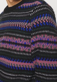 Neulepusero, jossa on musta pohja ja moniväriset kuviot vaaleanpunaisena, sinisenä, vihreänä ja valkoisena. Rikkaita pintarakenteita ja ribbireunus kauluksessa.