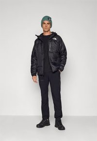 Juoda pūkinė striukė su užtrauktuku priekyje ir krūtinės kišene, derinama su juoda marškinėliais, juodomis kelnėmis ir turkio spalvos megzta kepure.