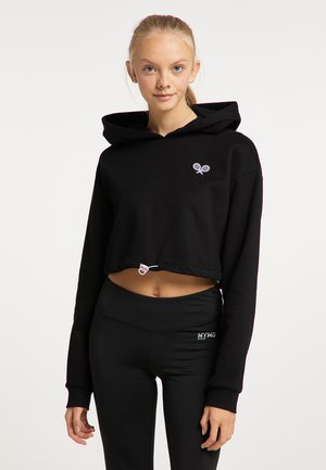 Jonge vrouw die een zwarte cropped hoodie met tennisracketslogo en zwarte hoge taille legging draagt, staand tegen een effen achtergrond.