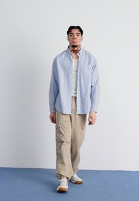 Chemise à rayures bleu clair et blanche à boutons, débardeur blanc, pantalon cargo beige et baskets blanches avec semelles beige. Debout sur une surface bleue.