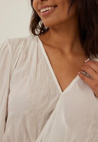 Weißes, strukturiertes Blusenshirt mit V-Ausschnitt und gekräuselten Schultern. Der Stoff wirkt leicht und leicht durchsichtig, mit einer glatten Oberfläche.