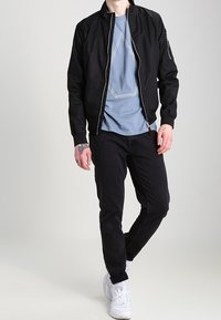 Svart bomberjacka med dragkedja, bärs över en ljusblå t-shirt med en vit geometrisk design, kombinerad med svarta jeans och vita sneakers.