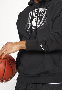 Czarna, kapturowa bluza z logo Brooklyn Nets z przodu, strukturalna tkanina, sznurki oraz biały logo Nike na mankiecie. Koszulka z piłką do koszykówki trzymaną w ręce.