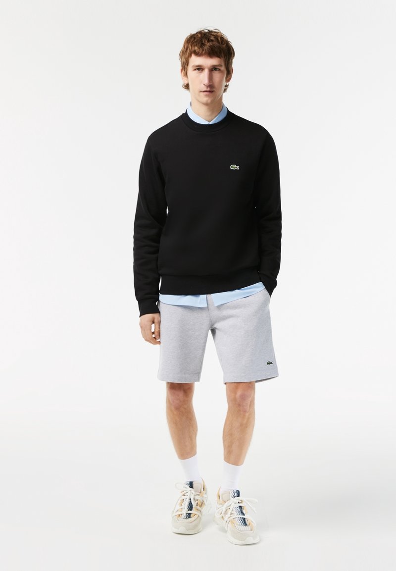 Jeune homme portant un sweat-shirt noir, une chemise bleu clair, un short gris, des chaussettes blanches et des baskets multicolores, debout sur fond blanc.