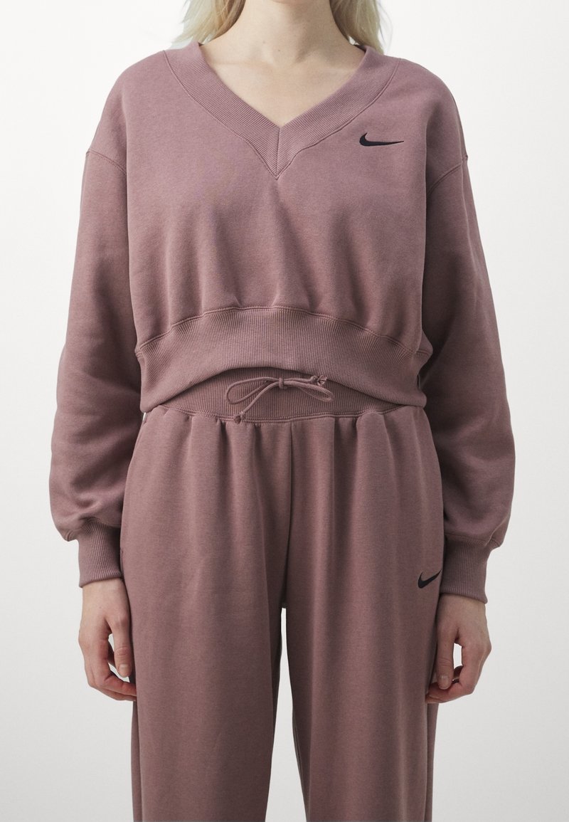 Ljus mauve sweatshirt och matchande byxor. Sweatshirten har V-ringning, ribbad mudd och byxorna har dragsko i midjan. Svart Nike-logotyp som accent.