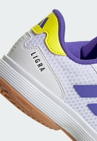 Chaussure de sport blanche avec des accents violets et jaunes, semelle en gomme, texte "LIGRA" sur le côté et "ADIWEAR" sur la bande de roulement de la semelle.