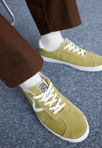 Green suede sneakers with white accents and laces. Round toe and rubber sole, featuring a logo tag on the side. Worn with white socks.