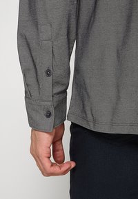 Grijze overhemden met visgraatpatroon met lange mouwen, voorzien van zwarte knopen bij de manchetten en een rechte zoom. Soepele textuur, casual ontwerp.