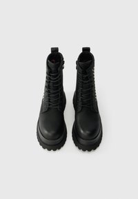 Fekete bőr bokacsizma vastag gumitalppal, fűzős kialakítással és díszstiftekkel az oldalukon. Simára csiszolt textúra, alacsony sarokkal.