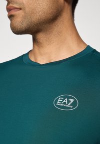 Tuva tuvumā redzams vīrieša kakls un krūtis, uz kuriem ir teal krāsas krekls ar baltu ovālu EA7 Emporio Armani logotipu kreisajā pusē.