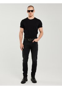 Bărbat purtând ochelari de soare negri, un tricou negru, blugi negri, o curea neagră și cizme negre, stând pe un fundal simplu și deschis la culoare.