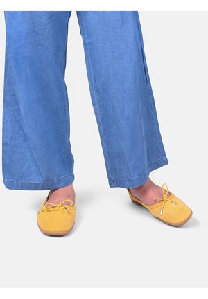 Persona che indossa pantaloni blu a gamba larga e ballerine gialle con un piccolo fiocco, in piedi su uno sfondo chiaro.