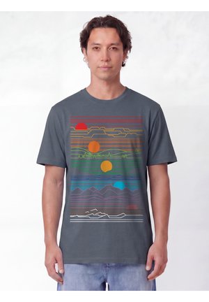 Homme portant un t-shirt gris avec des lignes horizontales colorées et des formes circulaires ressemblant à un paysage abstrait et des soleils.