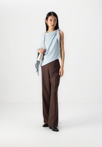 Top asimmetrico di colore blu chiaro con dettaglio nodo, abbinato a pantaloni larghi marroni e scarpe nere, in piedi su uno sfondo bianco semplice.