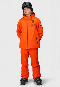 Παιδί που φορά φωτεινό πορτοκαλί μπουφάν και παντελόνι σκι, μαύρες μπότες σκι και μαύρα γυαλιά σκι στο μέτωπο, στέκεται με τα χέρια πίσω από την πλάτη.