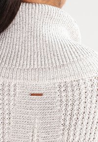 Sweter dzianinowy z wysokim, prążkowanym kołnierzem w jasnym beżu. Zawiera fakturowany wzór i małą skórzaną metkę z tyłu.