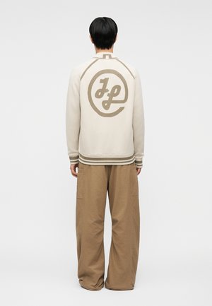 Person som står med ryggen mot, iført en kremfarget genser med en stor beige «Jp»-sirkel-logo på baksiden og løse, brune bukser.