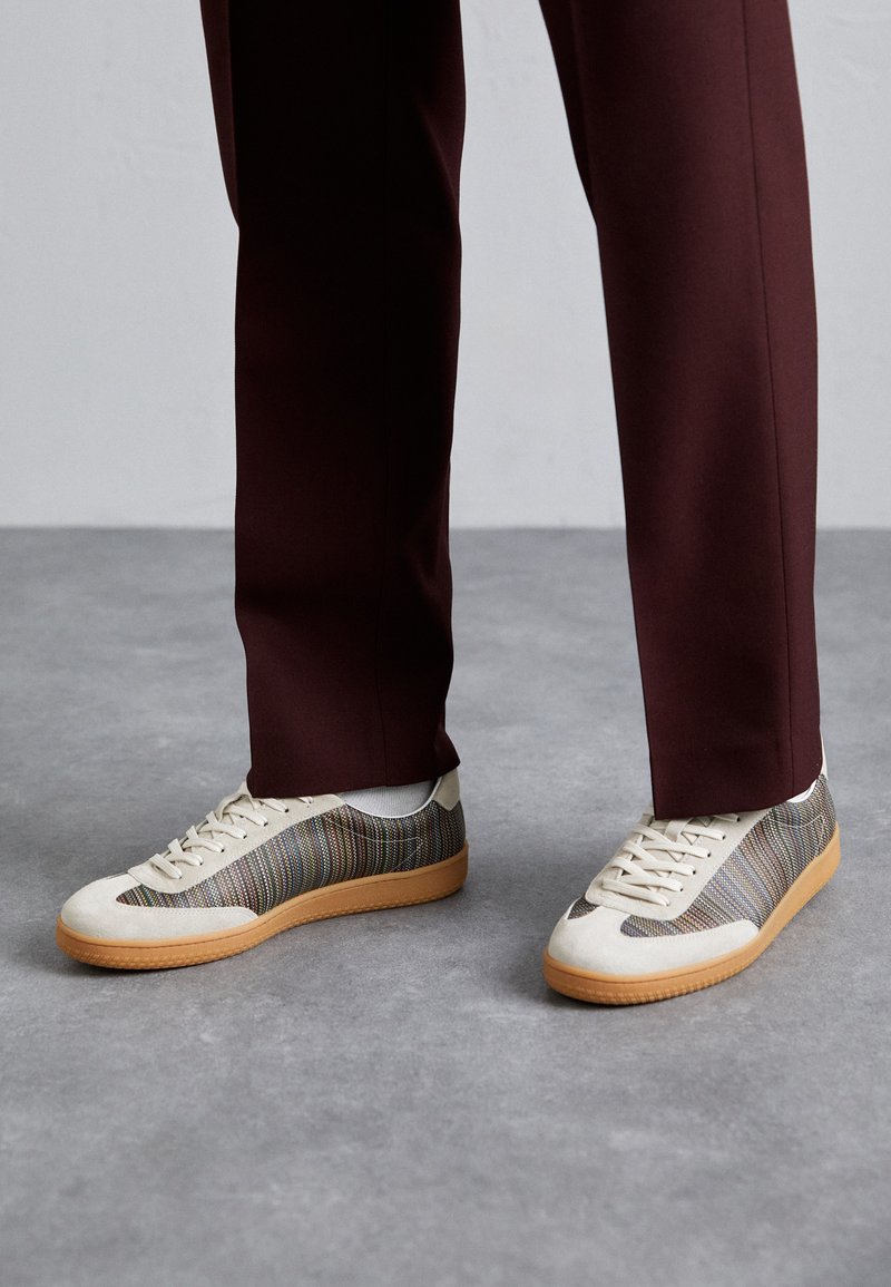 Baskets avec un dessus en toile beige présentant des rayures horizontales multicolores et une semelle en caoutchouc brun texturé. Associées à un pantalon bordeaux.