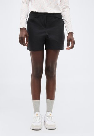 Fekete rövidnadrágot, hosszú ujjú, fehér mintás felsőt, világosszürke zoknit és fehér tornacipőt viselő személy áll egyszínű háttér előtt.