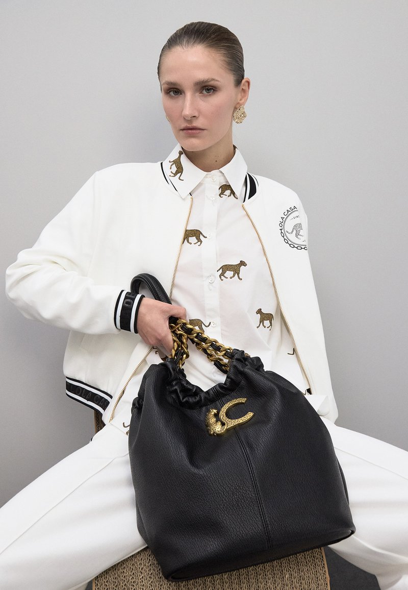 Bolso de cuero negro con una parte superior fruncida y un asa de cadena dorada, que presenta un emblema metálico. Modelo vistiendo una chaqueta bomber blanca y una camisa con estampado de leopardo.