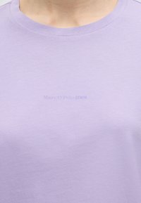 Κοντινό πλάνο ενός ατόμου που φοράει ανοιχτό μωβ μπλουζάκι με στρογγυλή λαιμόκοψη και αχνό κείμενο "Marc O’Polo DENIM" στο κέντρο του στήθους.