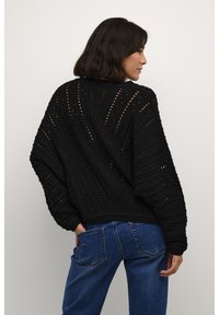 Svart stickad tröja med lös passform, med ett texturerat mönster och öppna detaljer. Matchad med blå denimjeans.