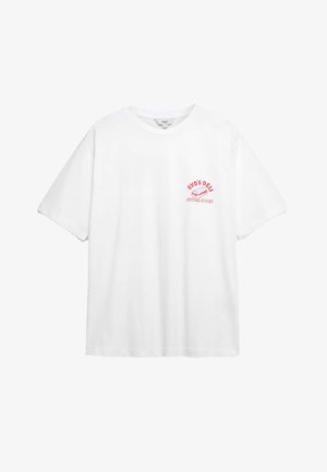 Camiseta de algodón blanca con mangas cortas y cuello redondo, que presenta un estampado gráfico en rosa en el lado izquierdo del pecho que dice "SYD'S DELI SUPER SUBS."