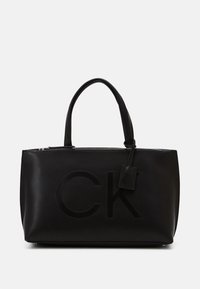 Sac tote en cuir noir avec fermeture éclair, mettant en avant un logo "CK" en relief, des poignées doubles et une étiquette amovible pour plus de détails.