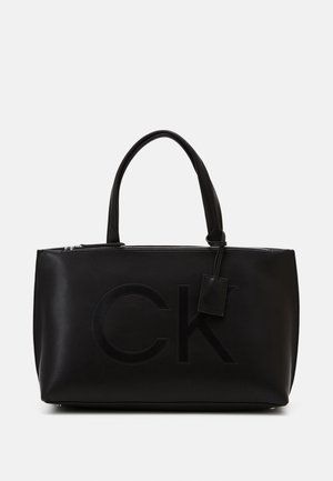 Black leather tote bag with zip closure, featuring an embossed "CK" logo, dual handles, and a detachable tag for added detail.