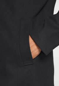 Abrigo negro con una textura suave, diseño estructurado, bolsillo lateral y un aspecto limpio y minimalista. La tela parece suave y cálida.