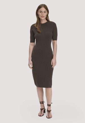 Frau trägt ein figurbetontes, knielanges, kurzärmeliges dunkelbraunes Kleid und passende, geschnürte Sandaletten mit Absatz, steht vor einem schlichten hellen Hintergrund.