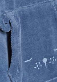 Blauer Plüschstoff mit einer weichen Textur, der eine Tasche in Form eines Katzenkopfes aufweist, versehen mit weißen Bestickungen für die Augen und die Nase.