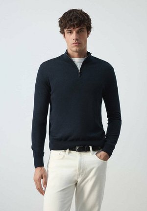Hombre con cabello oscuro y rizado, vestido con un suéter azul marino con medio cierre sobre una camisa blanca y pantalones blancos, de pie frente a un fondo claro y liso.