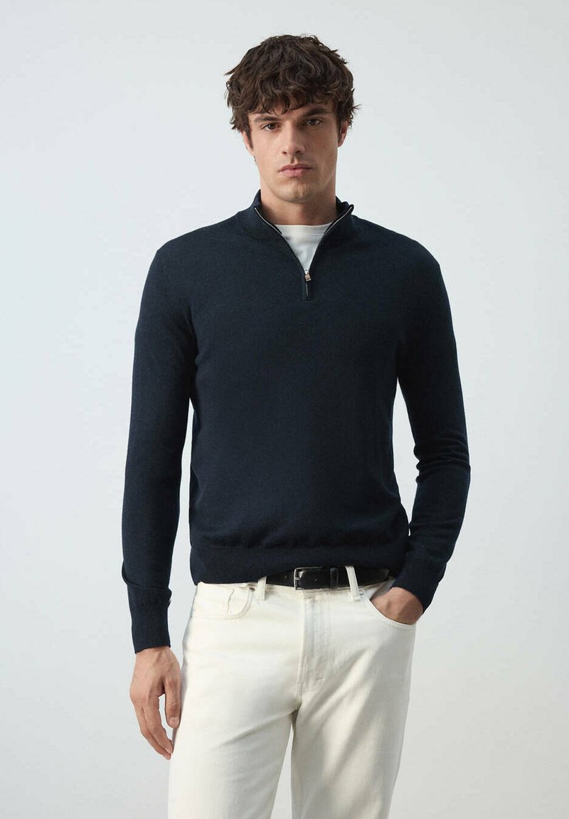 Homme aux cheveux bouclés foncés portant un pull bleu marine à demi-fermeture éclair par-dessus une chemise blanche et un pantalon blanc, debout devant un fond clair uni.