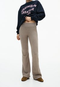 Vrouw in beige gebreide flared broeken, bruine instap schoenen en een marineblauwe sweatshirt met de tekst "Birmingham Creative", staand tegen een witte achtergrond.